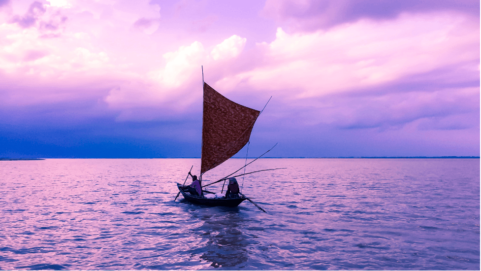 Two people in a small wooden sailboat with a brown sail, gliding across calm water under a purple and pink sunset sky.