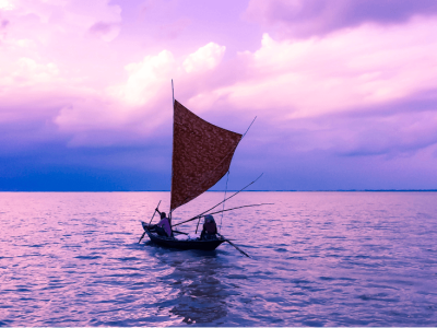 Two people in a small wooden sailboat with a brown sail, gliding across calm water under a purple and pink sunset sky.